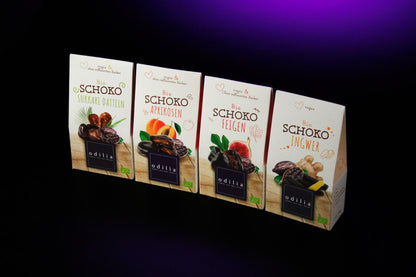 Die 100g Packungen der Bio-Schoko-Aprikosen, Bio-Schoko-Ingwer, Bio-Schoko-Feigen und Bio-Schoko-Sukkari-Datteln. Die Verpackungen zeigen die jeweiligen Trockenfrüchte und Zutaten und machen Lust, die leckeren Schokoladen-Kreationen zu probieren.